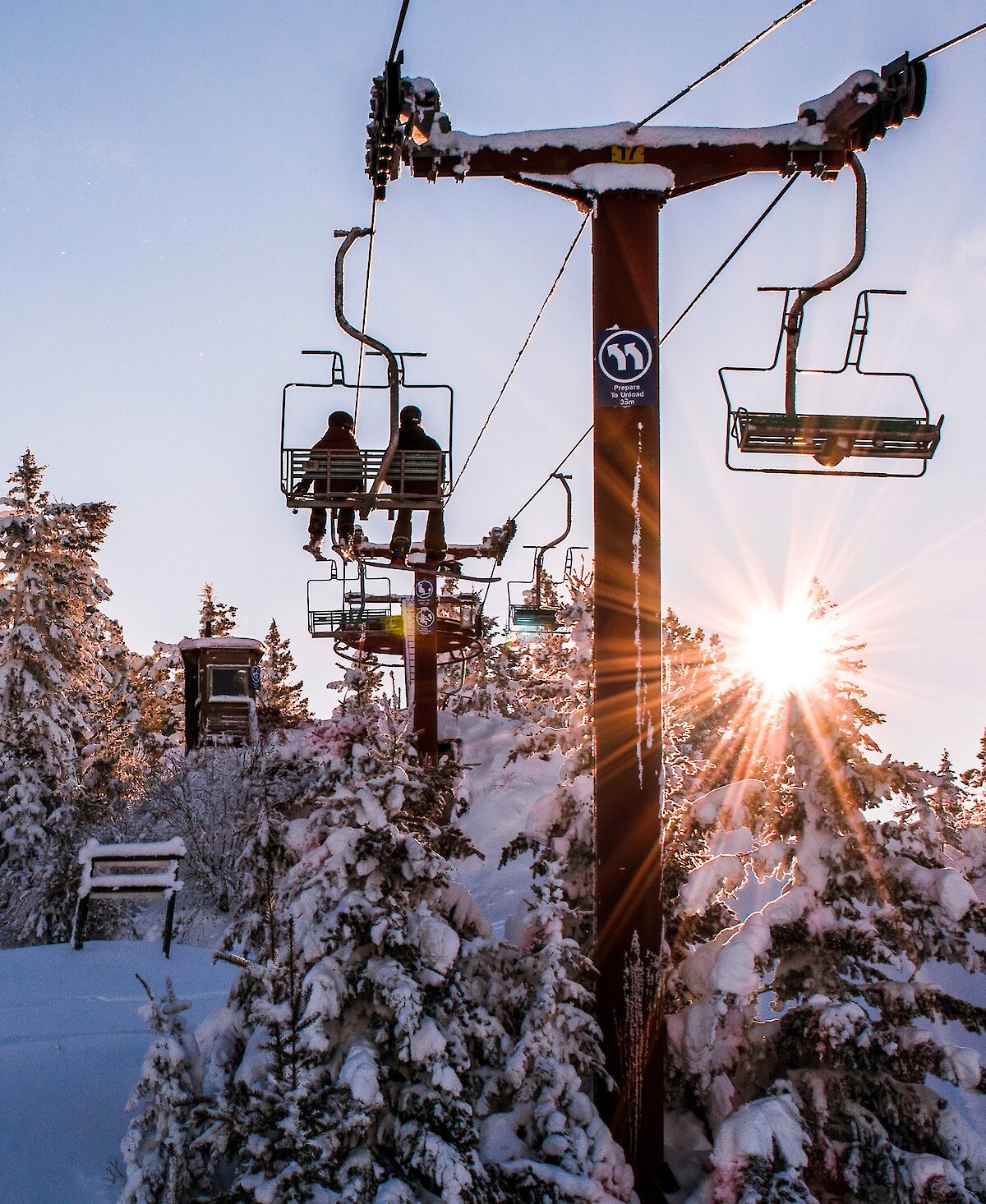 A skier and boarder riding the chairlift at Harper Mountain Ski resort with the Sun poking through the snow-covered trees.