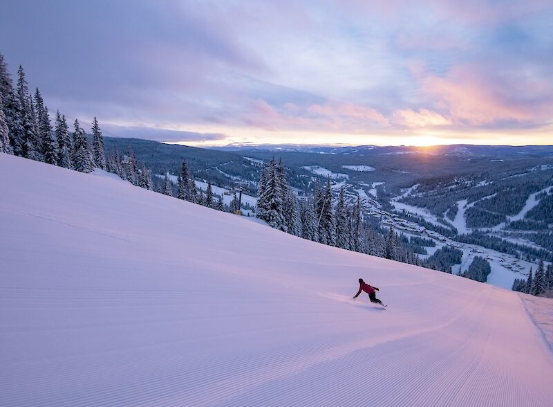 Snowboarding carving down a pristinely groomed trail at Sun Peaks Resort with a view of the mountain's ski trails and sunset in ithe background.