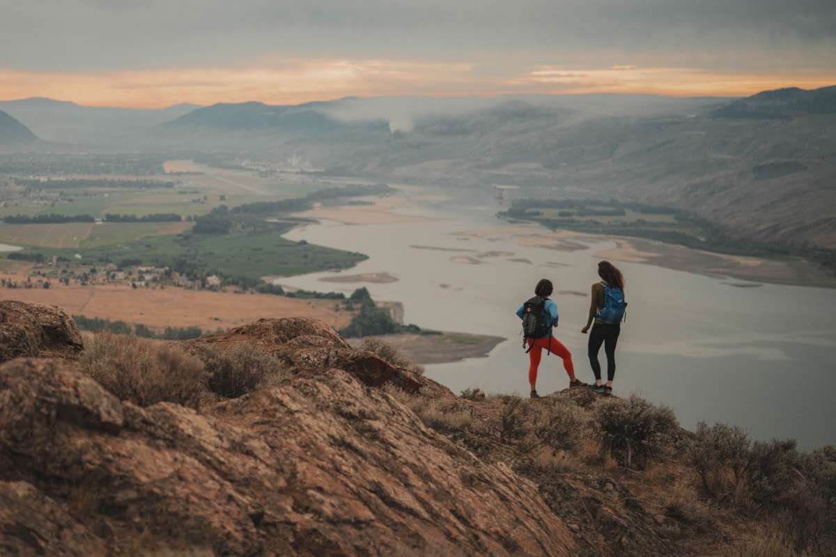 Two young women hike at the top of battle bluff. You can see the Thompson River surrounded by mountains and green fields