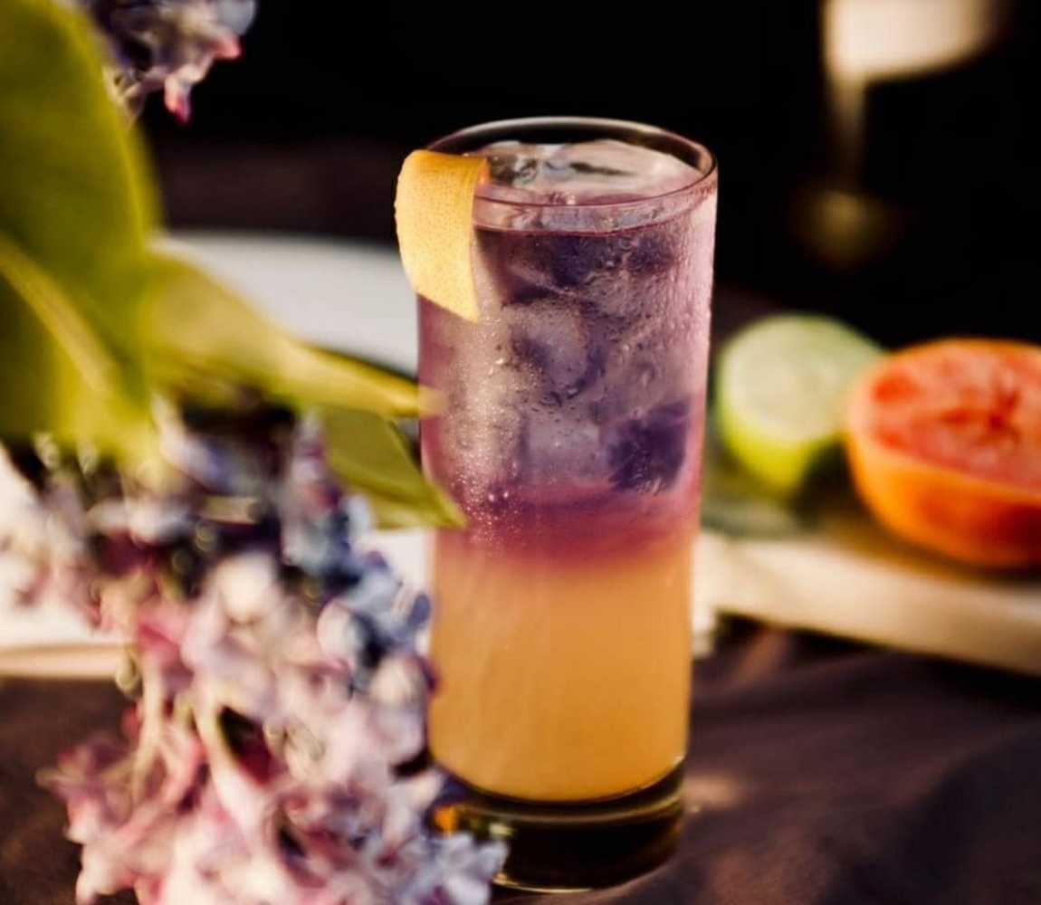 a purple and orange cocktail in a tall glass set amongst a flower display