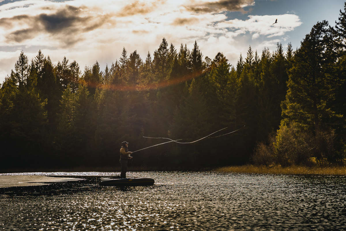 Man casting from an inflatable boat with the sun shining and forest in the background in Kamloops, BC.