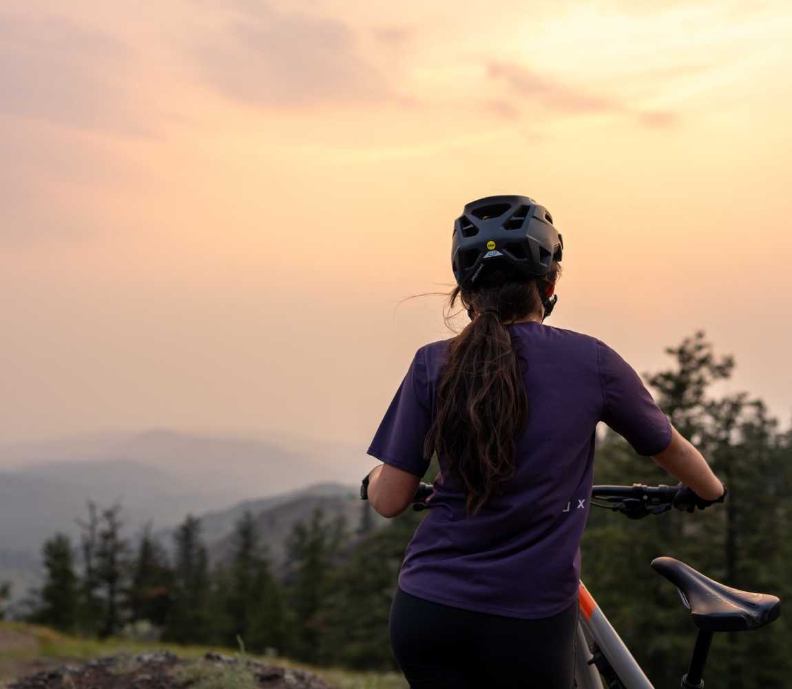 A woman holding her mountain bike with pine trees, a mountain, and sunset in the background in Kamloops, BC.