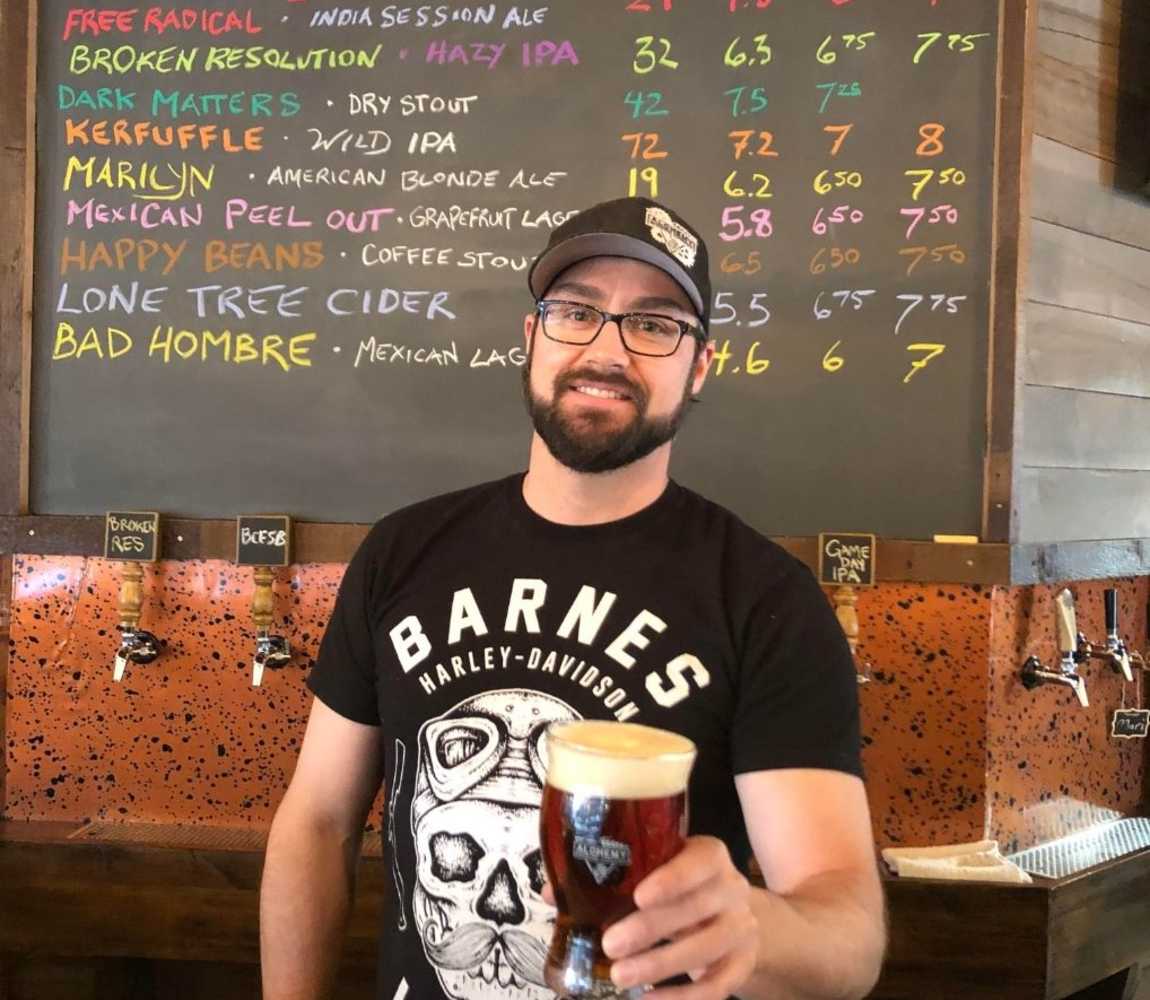 Alchemy Brewing Company brewer, Al Renner, standing in front of their tap list holding a beer