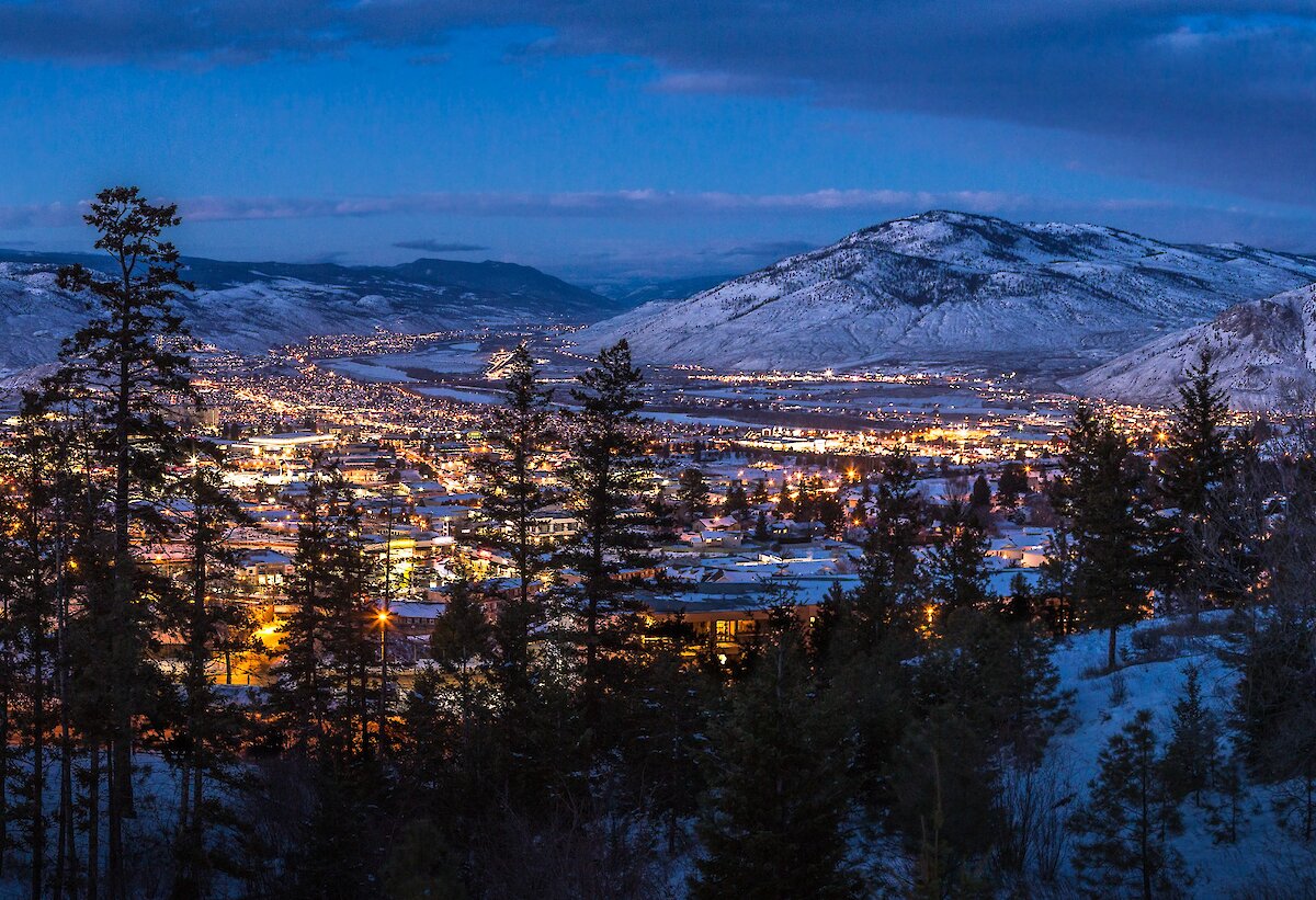 A view of Kamloops at night, with snowy hills in the distance and foreground, and the city lit up with many bright lights.