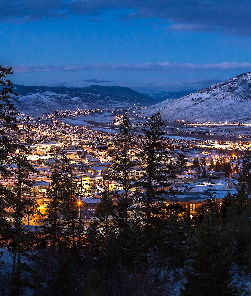 A view of Kamloops at night, with snowy hills in the distance and foreground, and the city lit up with many bright lights.