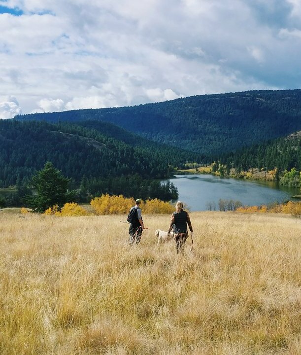 A couple and their dog hike through the Lac du Bois Grasslands near Kamloops. Fall colours are visible on the trees in the distance, and a calm lake reflects a blue sky.