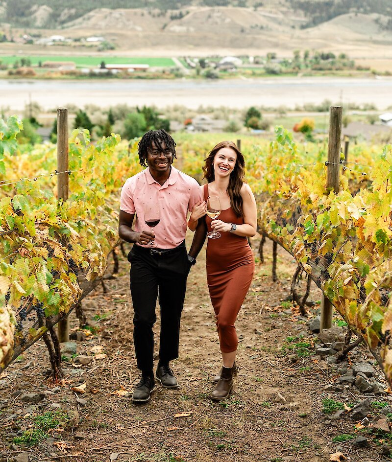 Couple walking through the fall coloured vines with glasses of wine at Sagewood Winery in Kamloops, BC.