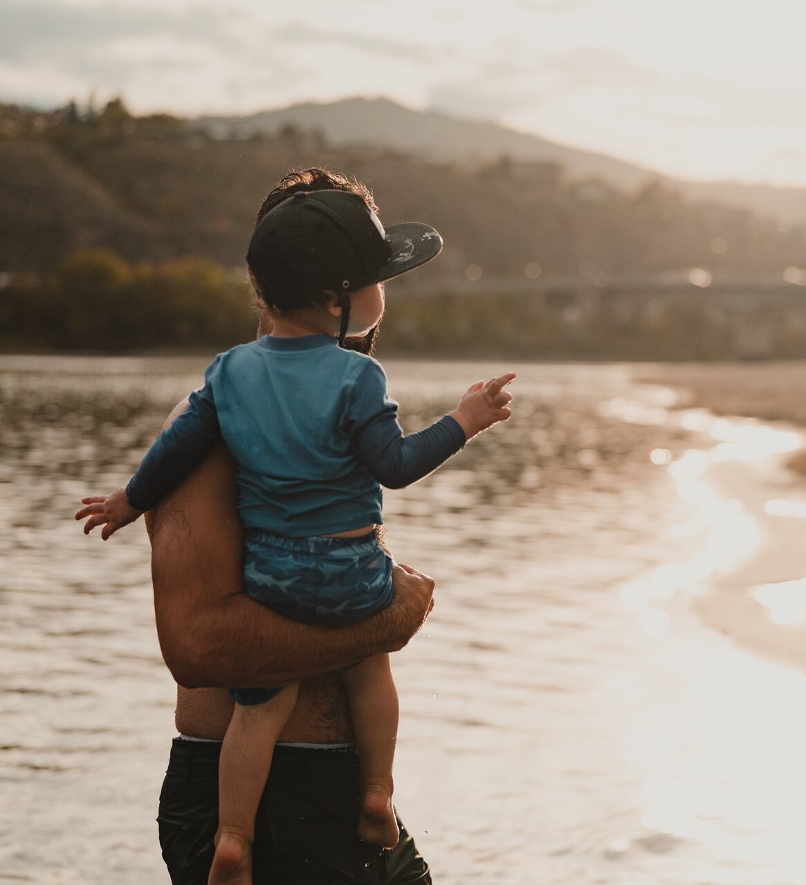 A man and child enjoy the beach at Overlander Park in Kamloops, standing in the water near the sand, with the sun setting behind a bridge in the background.