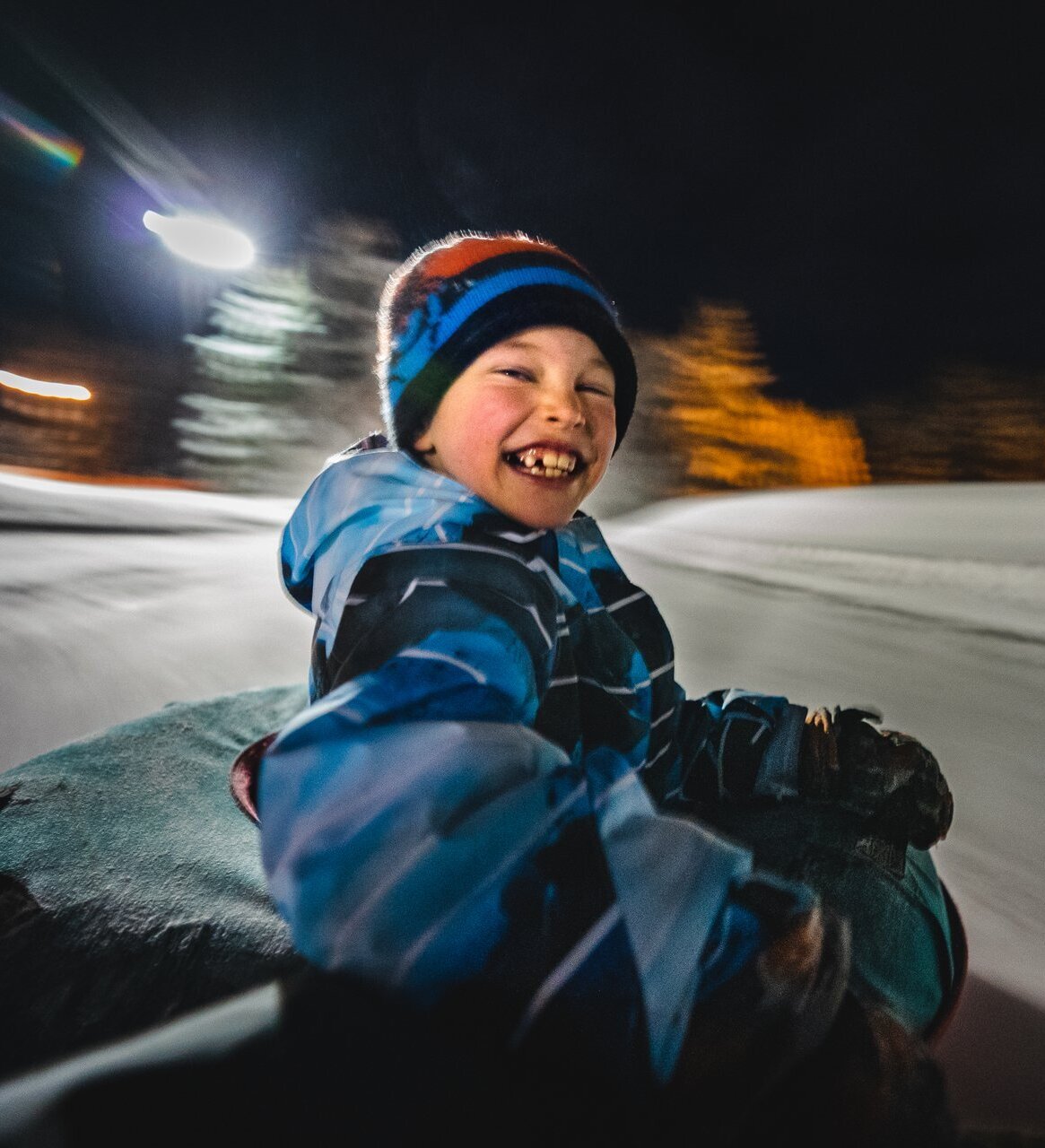 A child in winter gear grins at the camera while riding a tube down a snowy hill. The background is blurred indicating high speed.