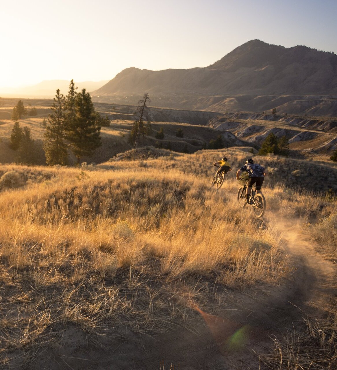 Two people ride mountain bikes through grassland hills with the sun setting in the distance.