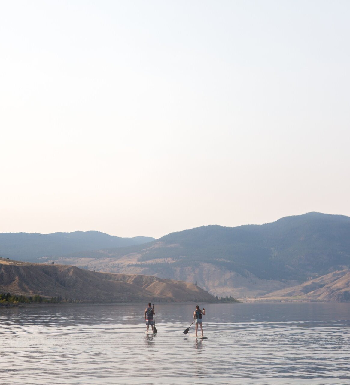 Two people paddleboard on a smooth lake, with hills in the distance and a clear sky above.