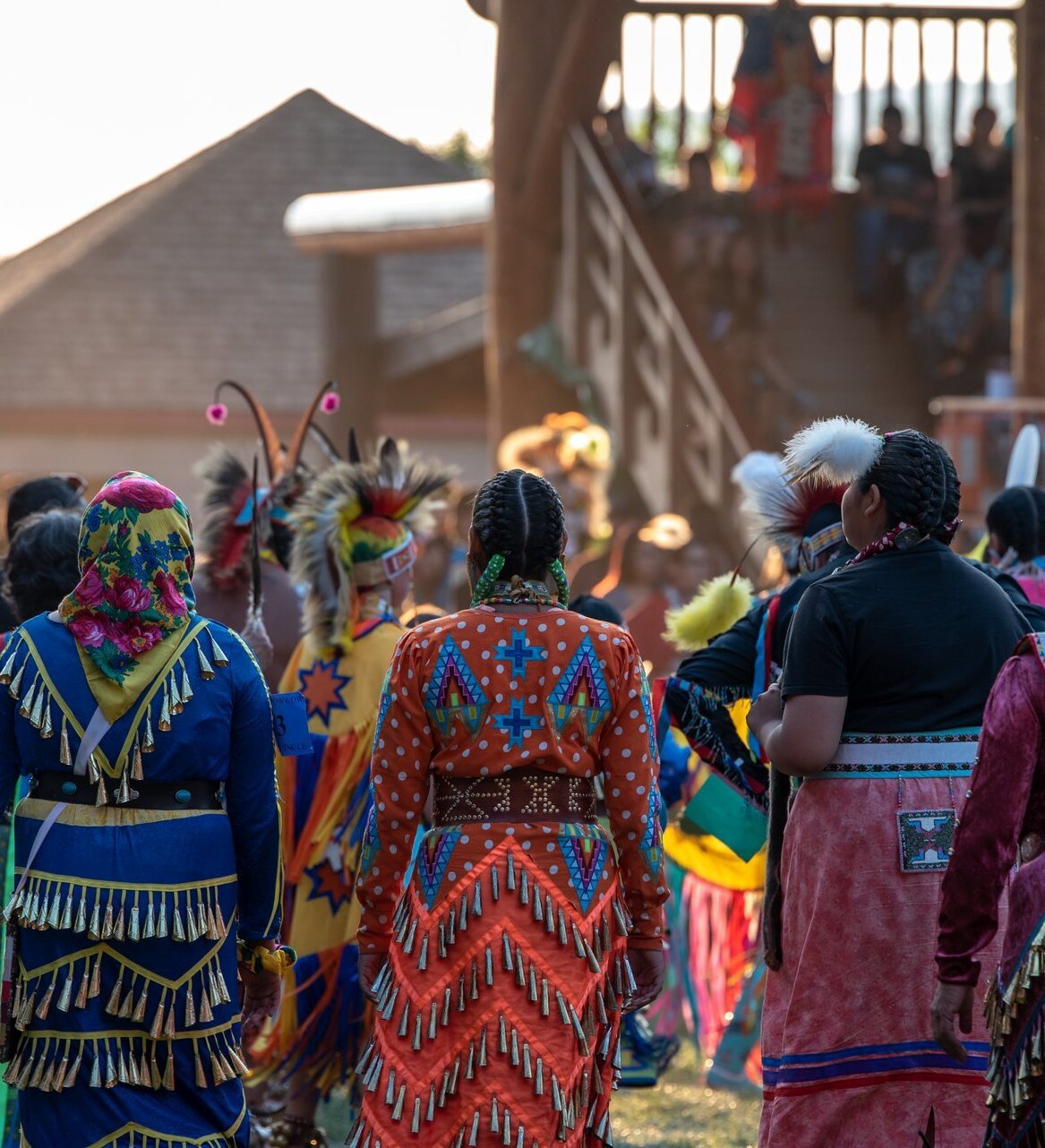 A group of women in traditional secwépemc dress approach the powwow arbour with a crowd of attendees.