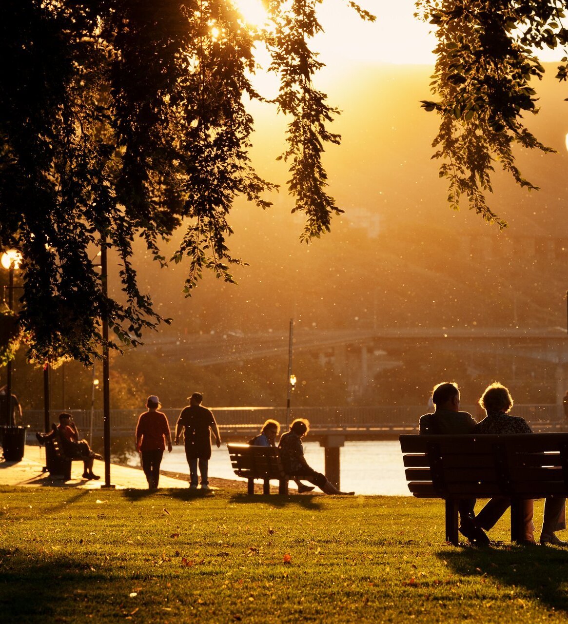 People sit on benches and walk through a park along the river. The path is lined with lamp posts. The sun is setting in the background, creating a lot of golden light.