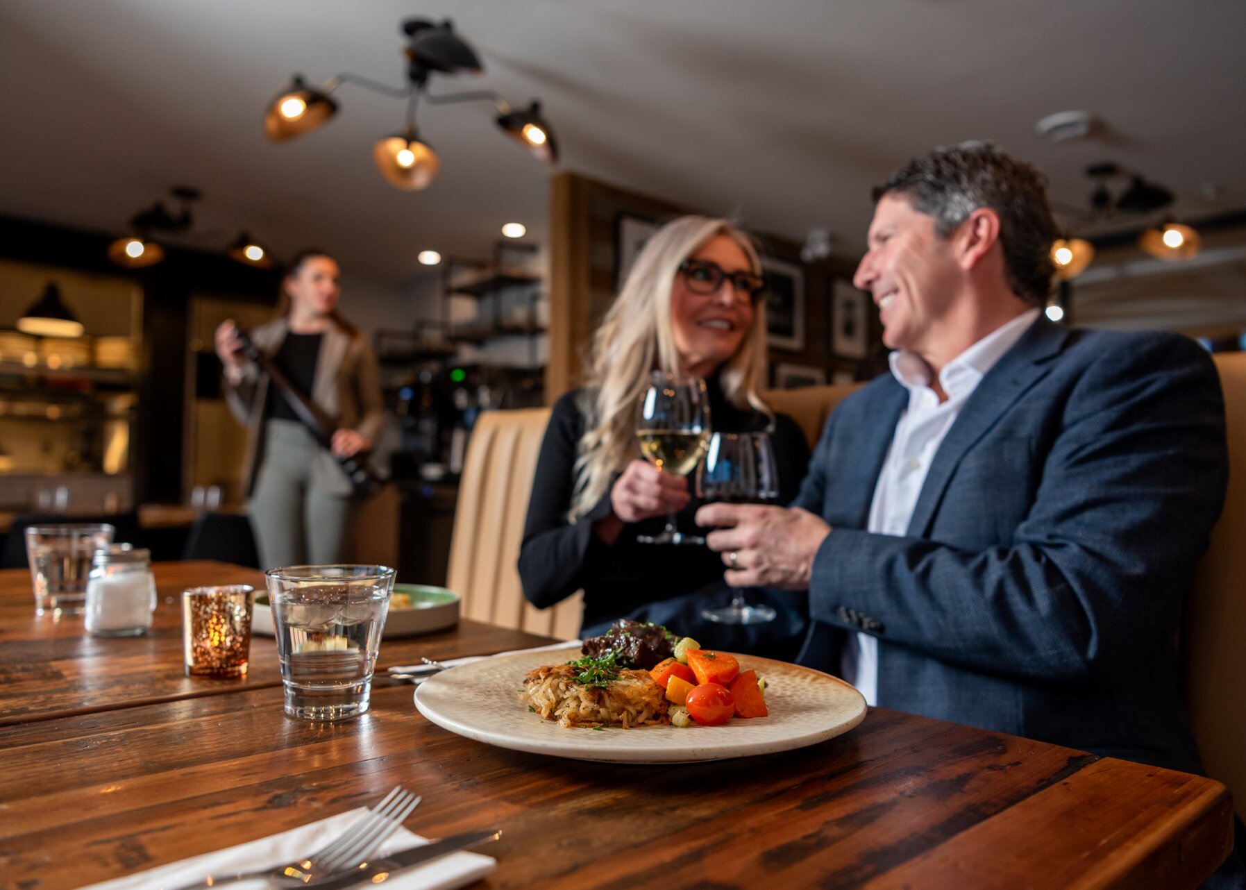 A couple cheers their wine glasses with delicious entrees in front of them at Isagani Farm-to-Table in Kamloops, BC.