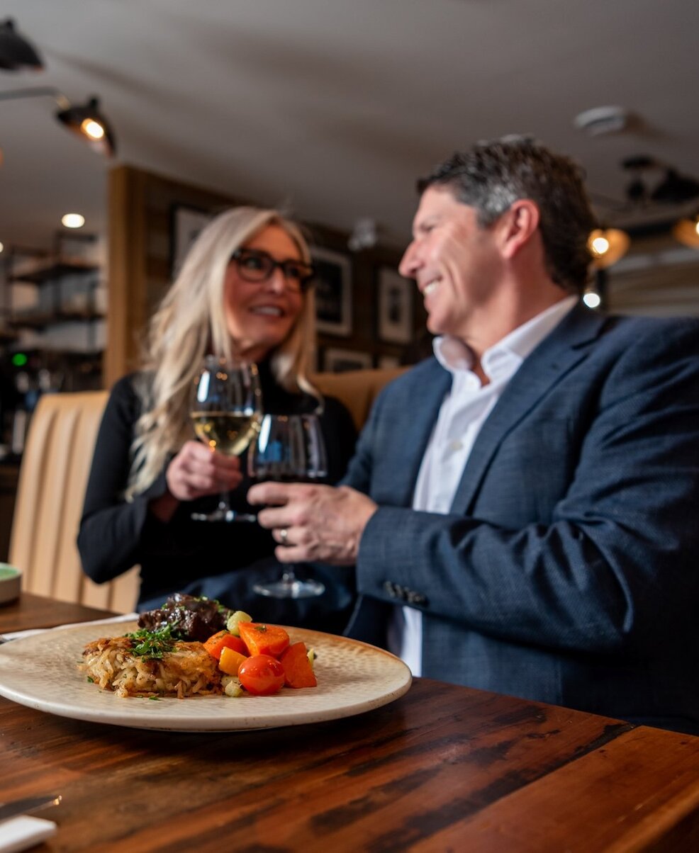 Couple cheers-ing their wine glasses over their delicious dishes at Isagani Farm-to-Table located in Kamloops, BC.