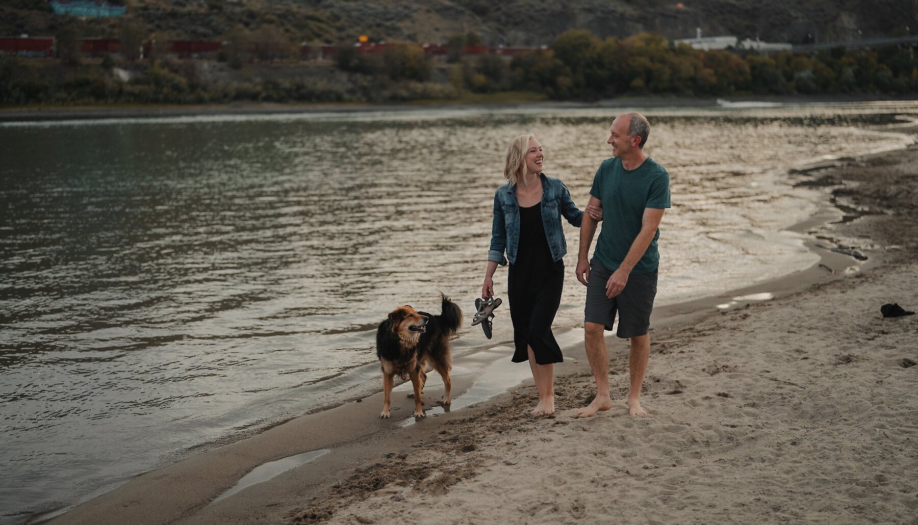 A couple walking their dog along the Thompson River shoreline at Overlander Park Beach in Kamloops, BC.