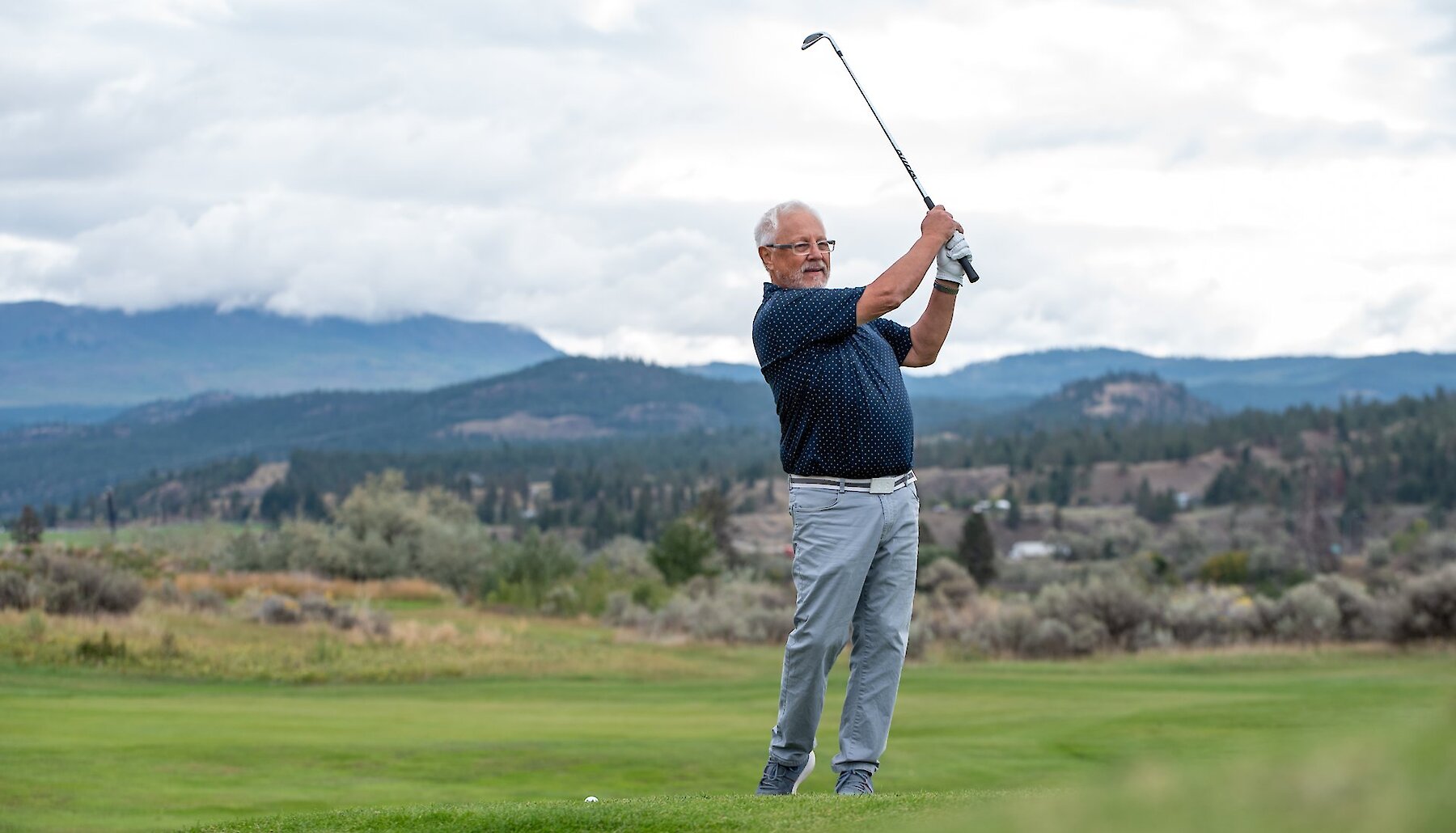 A golfer swinging his club with a mountain landscape behind him in Kamloops, BC.