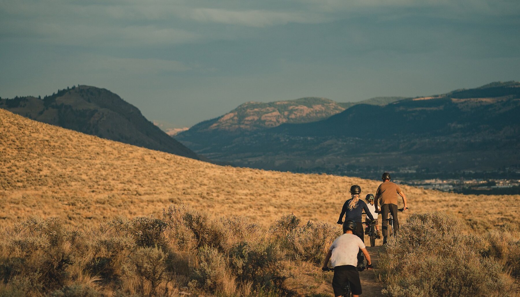 A family mountain biking through the Lac du Bois Grasslands with the mountain range in the background in Kamloops, BC.