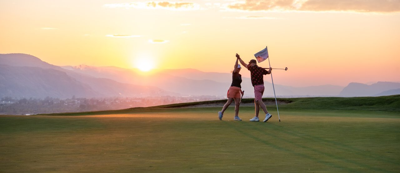 Two golfers high five on one of Kamloops' golf courses. The sun is low behind them and sunset blues, pinks and golds paint the hills and city in the distance.