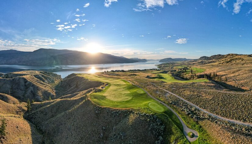 Aerial shot of Tobiano – an award-winning golf course in Kamloops BC. Golf greens and fairways are interspersed with arid grasslands under a blue sky, and the sun is setting over Kamloops Lake in the distance.