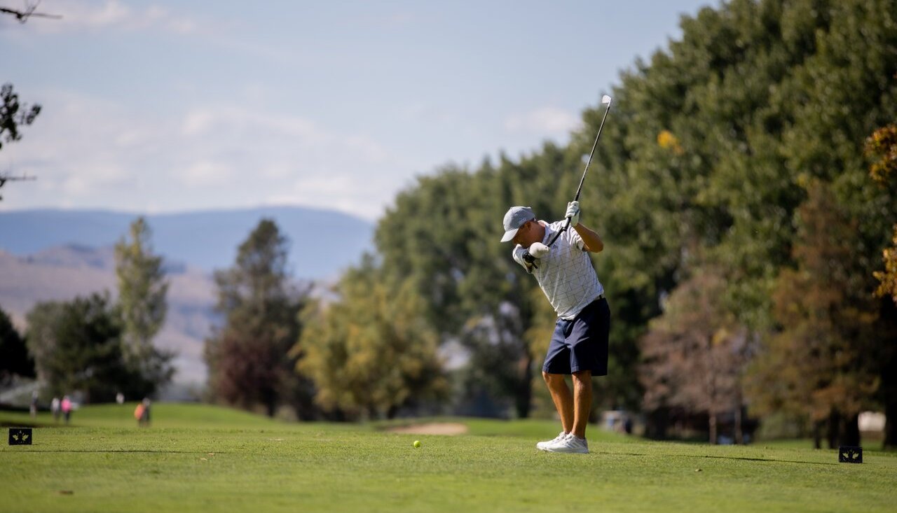 A man tees off at the Kamloops Golf & Country Club, with a line of trees on the right and blue skies above.