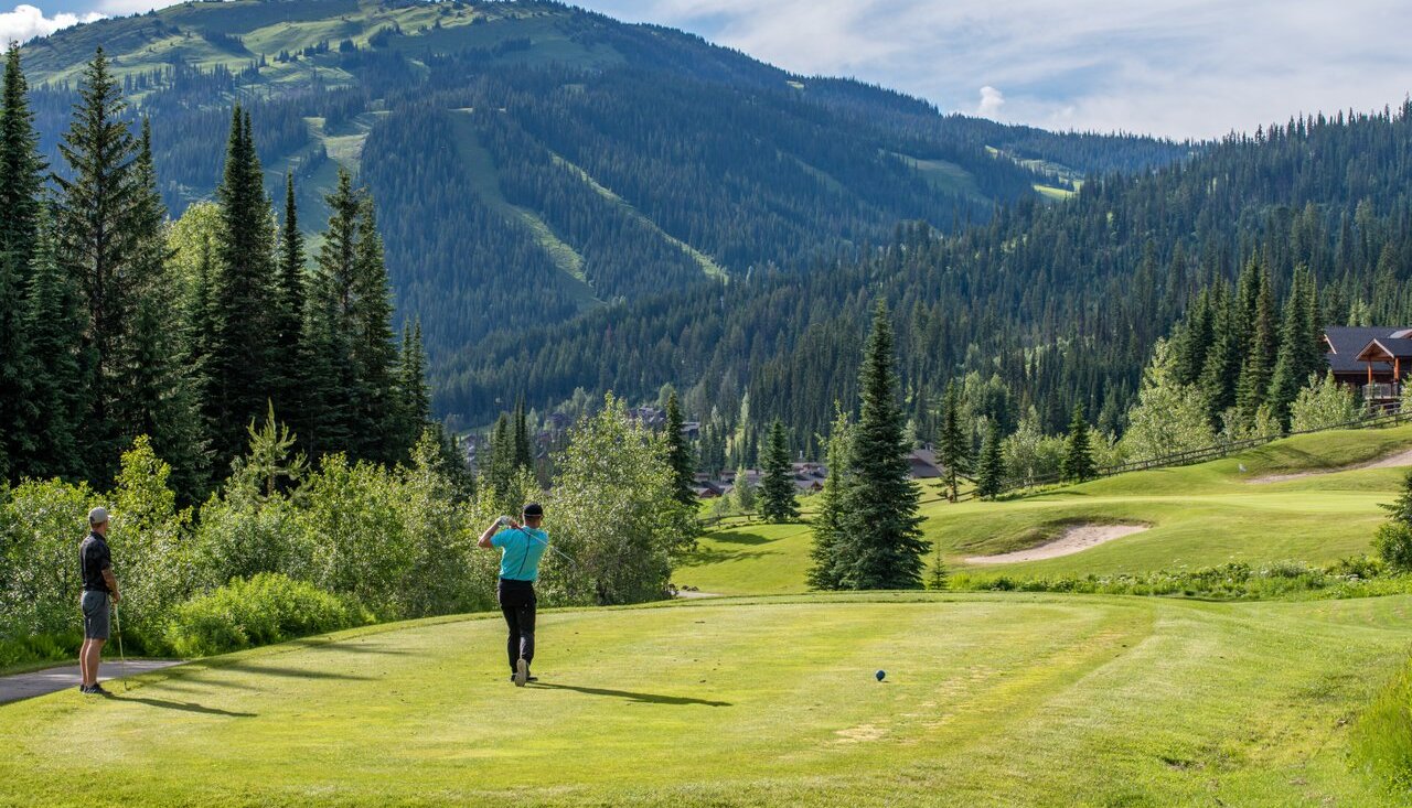 A man golfs at Sun Peaks BC. He is standing on a fairway looking out over a sloping course with sand traps and rising mountains in the distance.