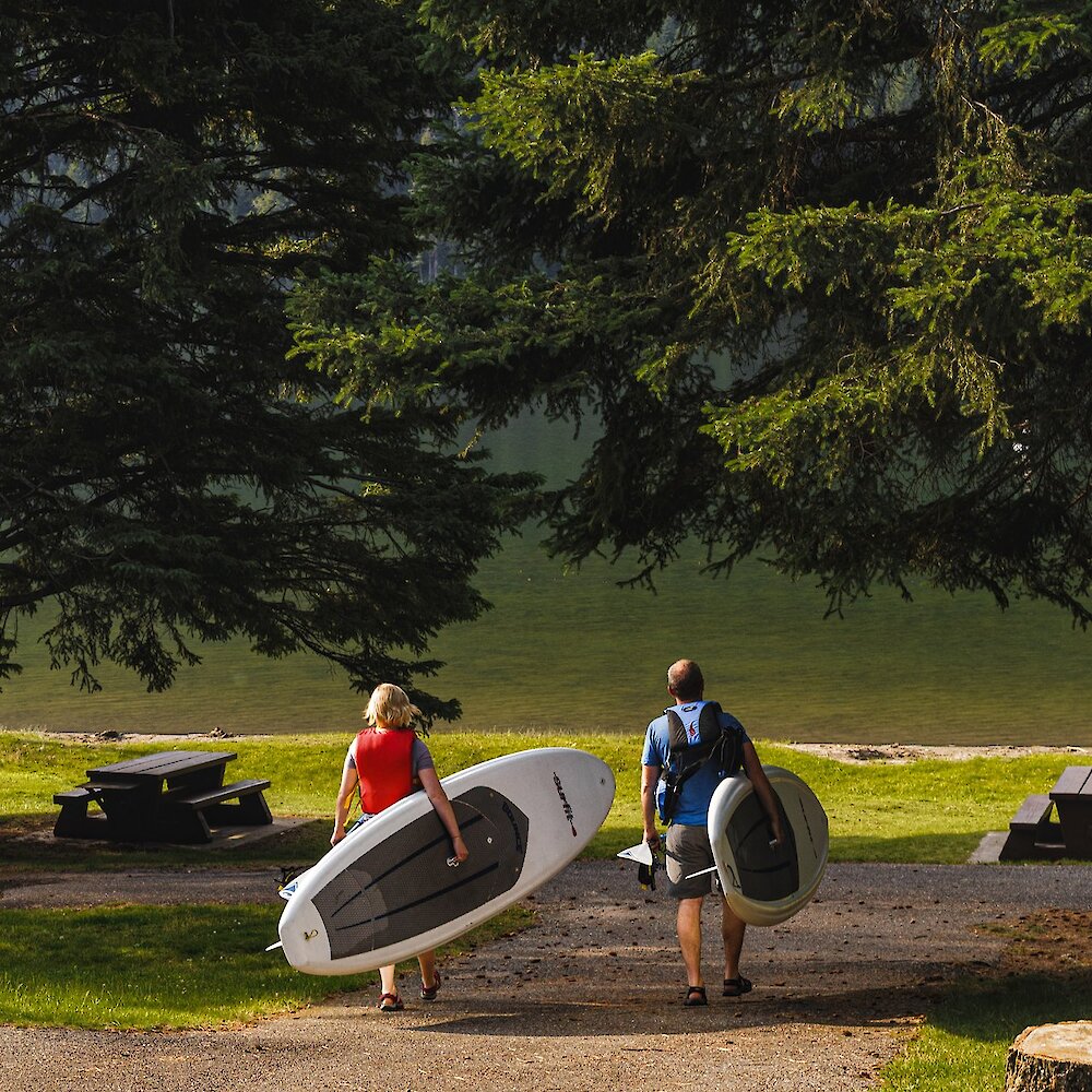 A couple carrying their paddleboards down to Paul Lake to launch onto the water.