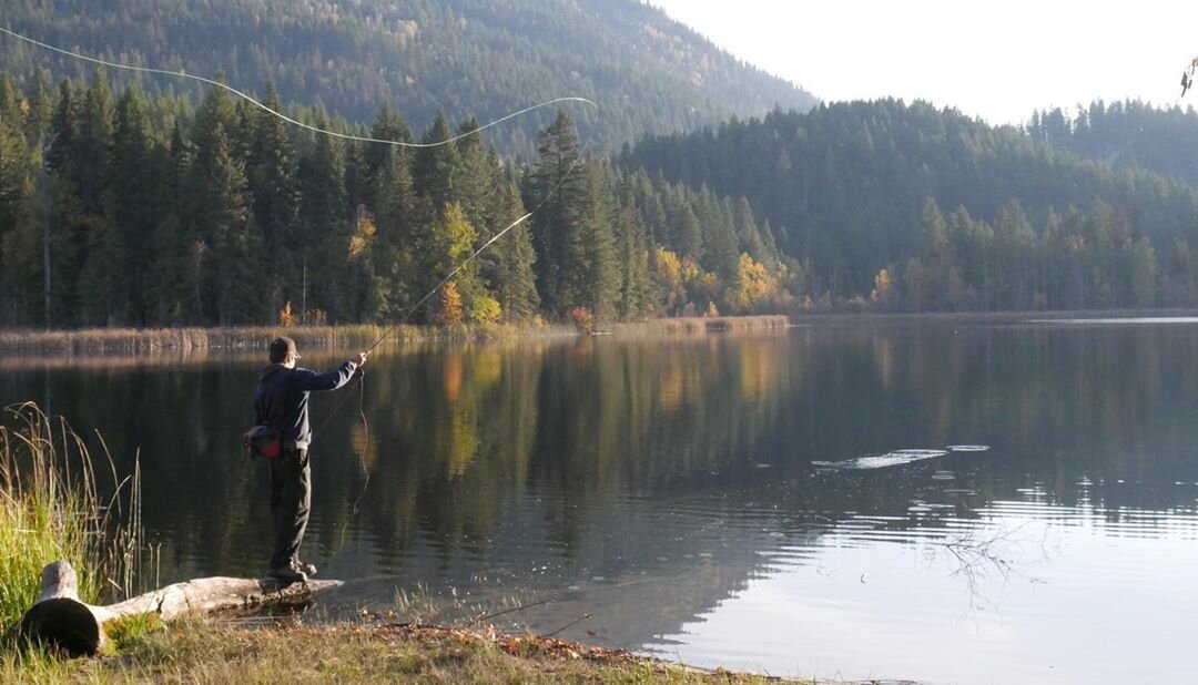 A fisher casts into calm lake, with autumn colours lining the shore behind them.