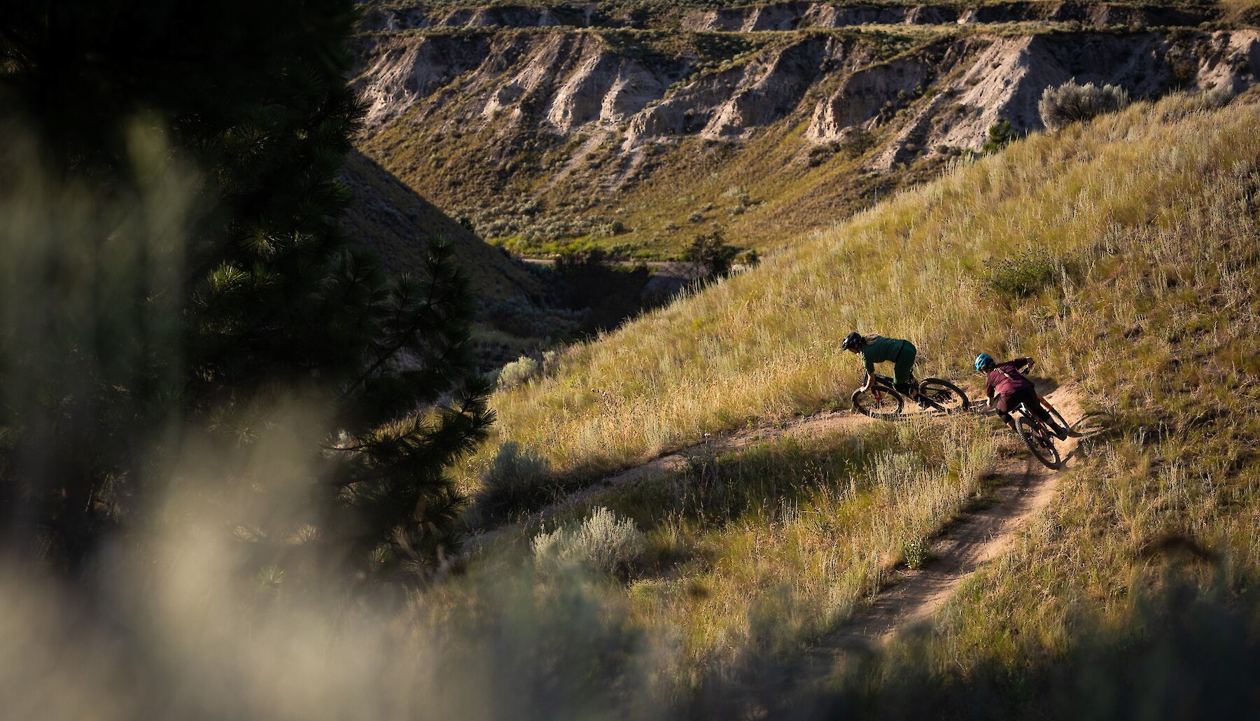 A mountain biker is seen traversing a trail through an open field at high elevation. Low clouds obscure part of the valley below.