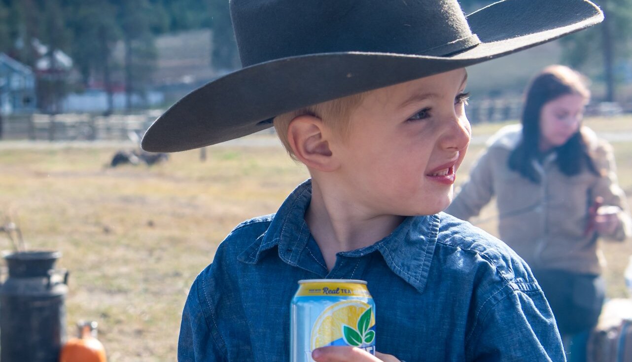 A young boy in a cowboy hat, denim, and rodeo belt enjoys some juice at an outdoor event.