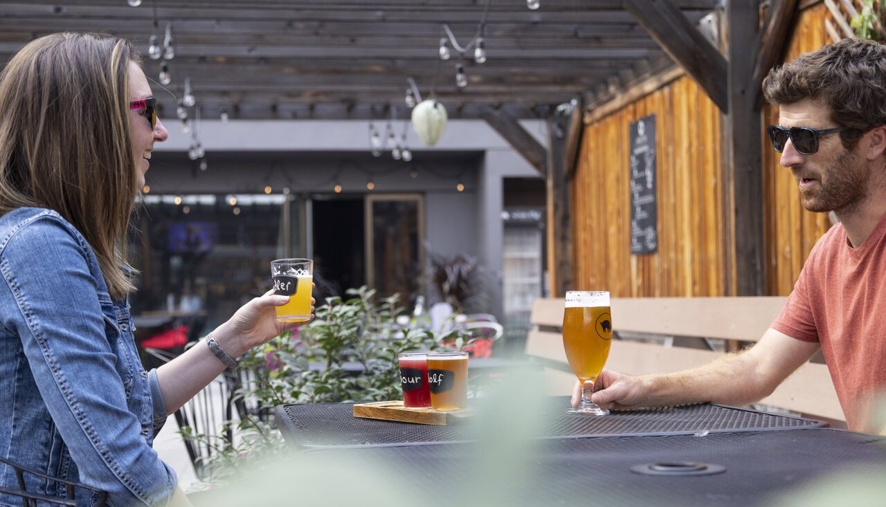 Two friends drink beer at the Noble Pig in Kamloops, British Columbia. They sit on a patio with lights strung overhead.