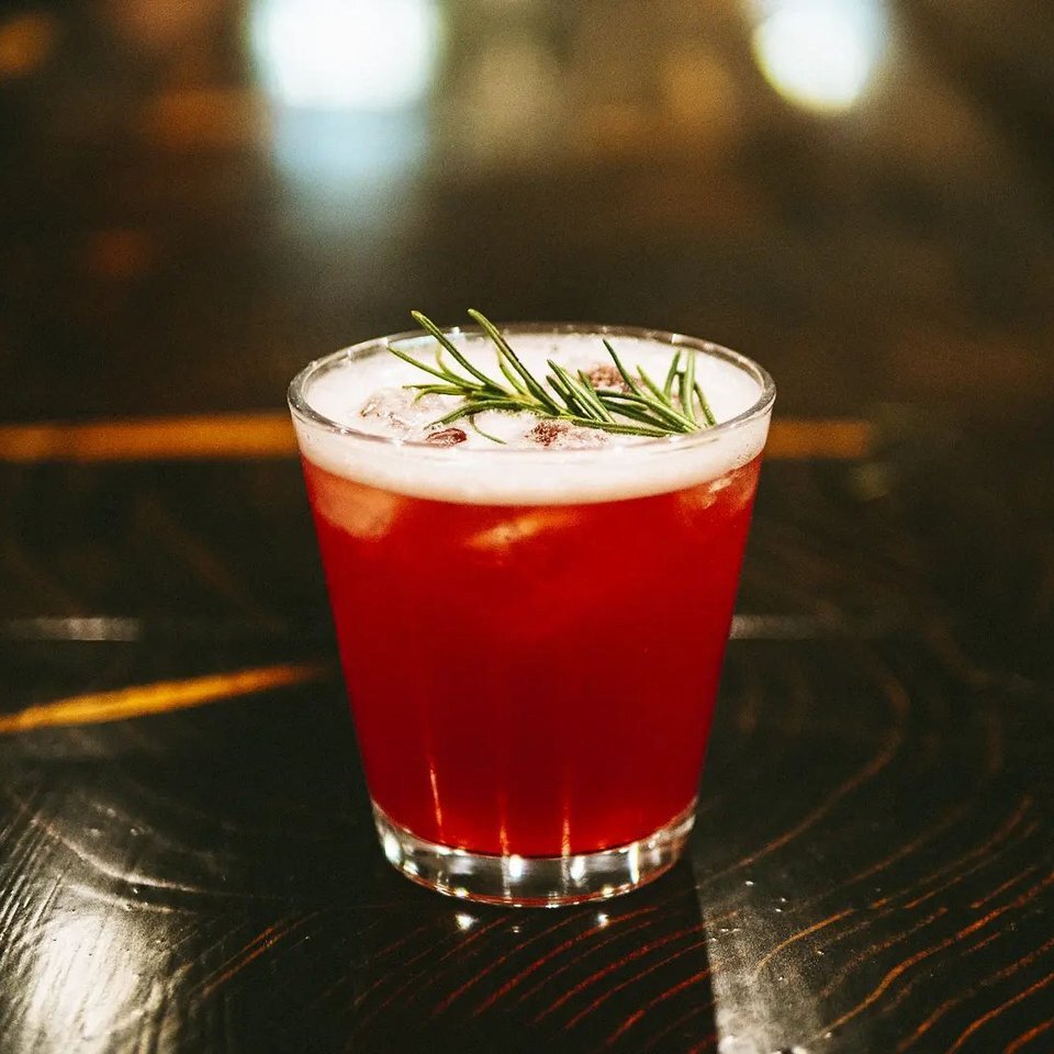 A gin drink sits on a dark bar at Kamloops BC's Gin Lane Distillery. It's a glowing red and garnished with a sprig of rosemary.