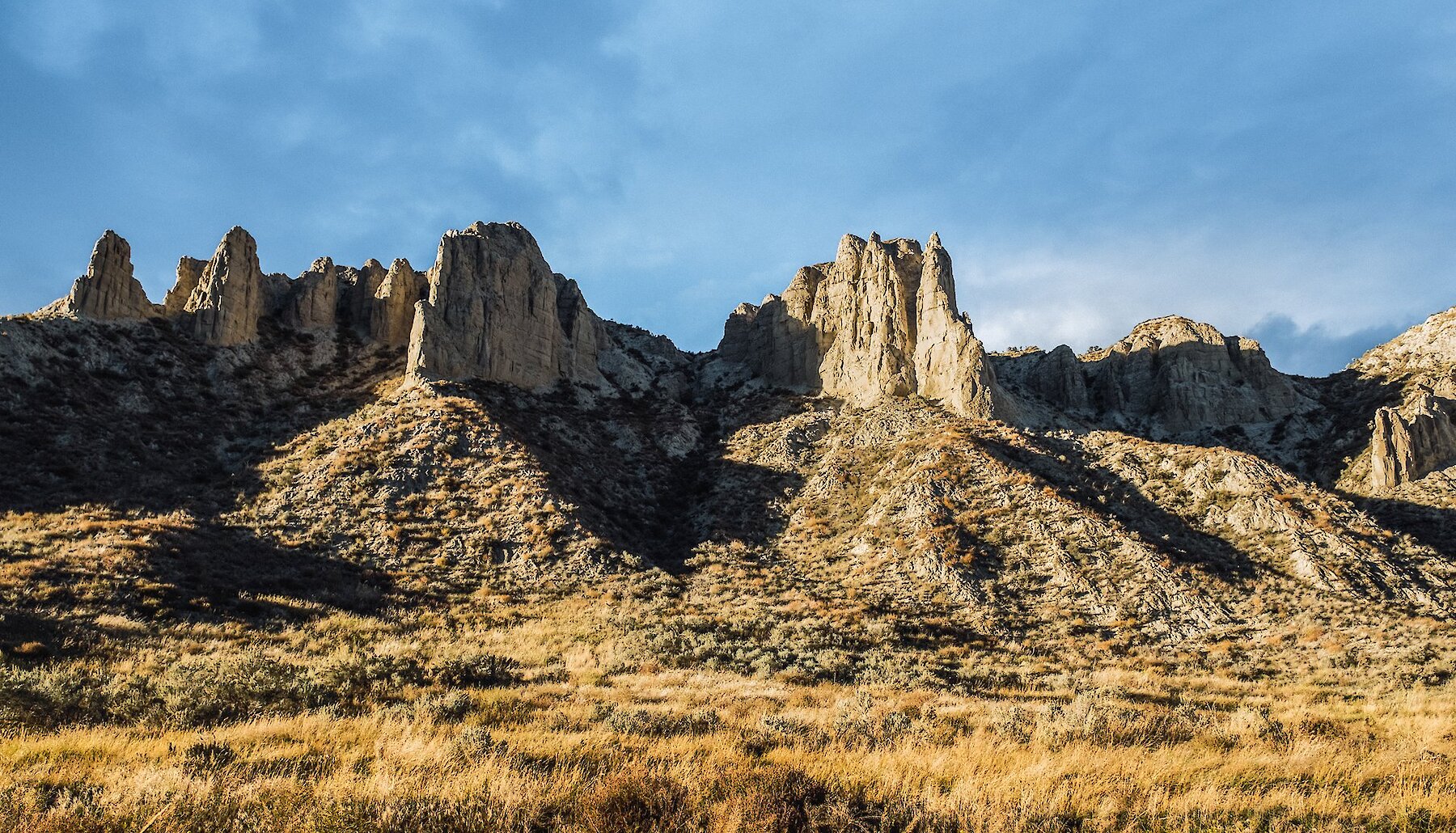 A dramatic ridge of hoodoo formations against a bright blue sky, with golden grasslands and sagebrush hills below.