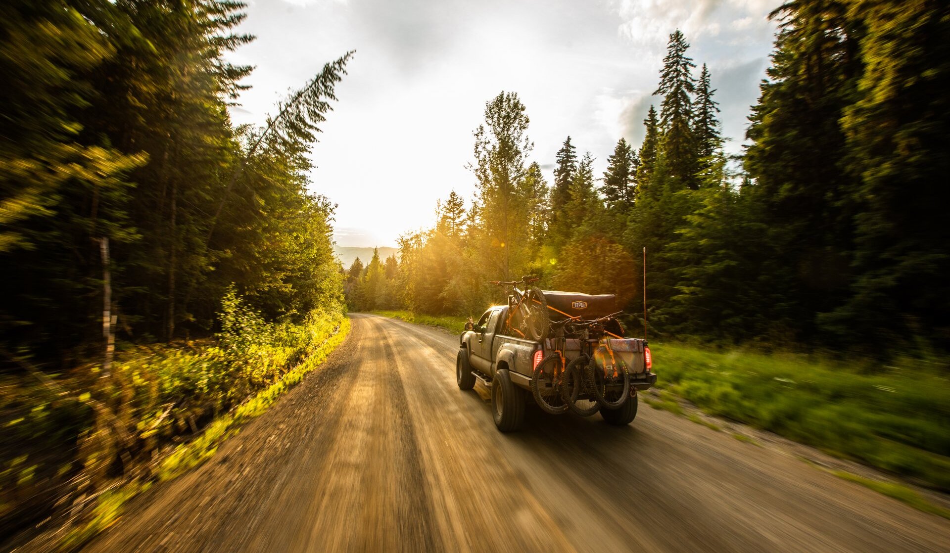 A truck loaded with mountain bikes drives up a forest road towards a sunset