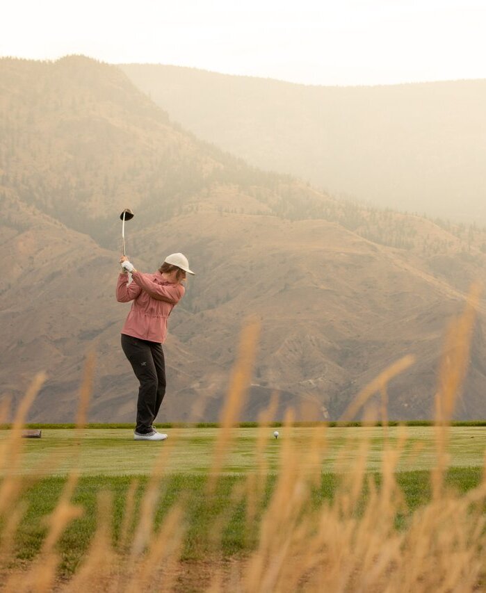 Two golfs prepare to tee off on a smooth green at Tobiano. There is tall wild grass in the foreground, and low sun on the hills in the distance.