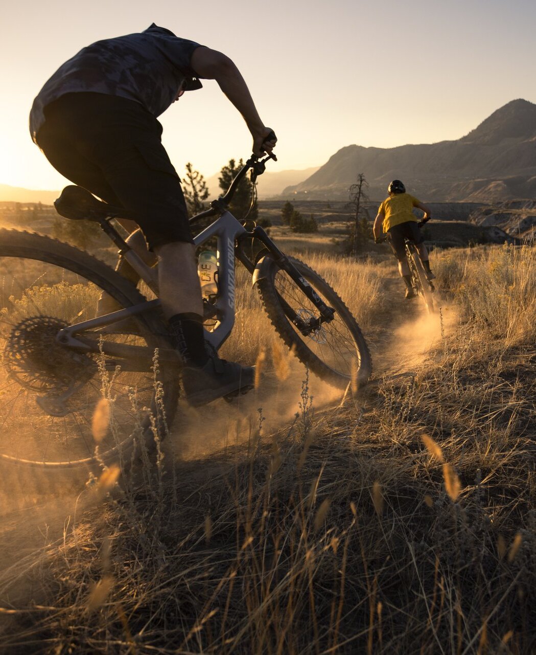 Two mountain bikers ride a grasslands trail in Kamloops. Hoodoos and sandstone bluffs are visible in the distance.