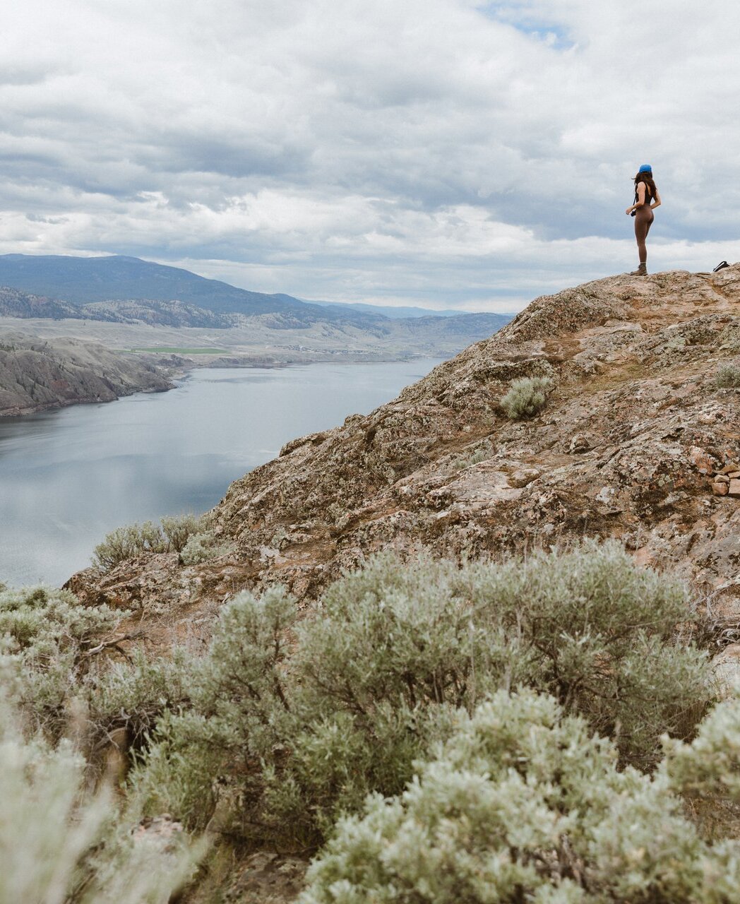 A young female hiker stands on a bluff overlooking a calm lake, with rolling grassland hills in the distance and sagebrush in the foreground.