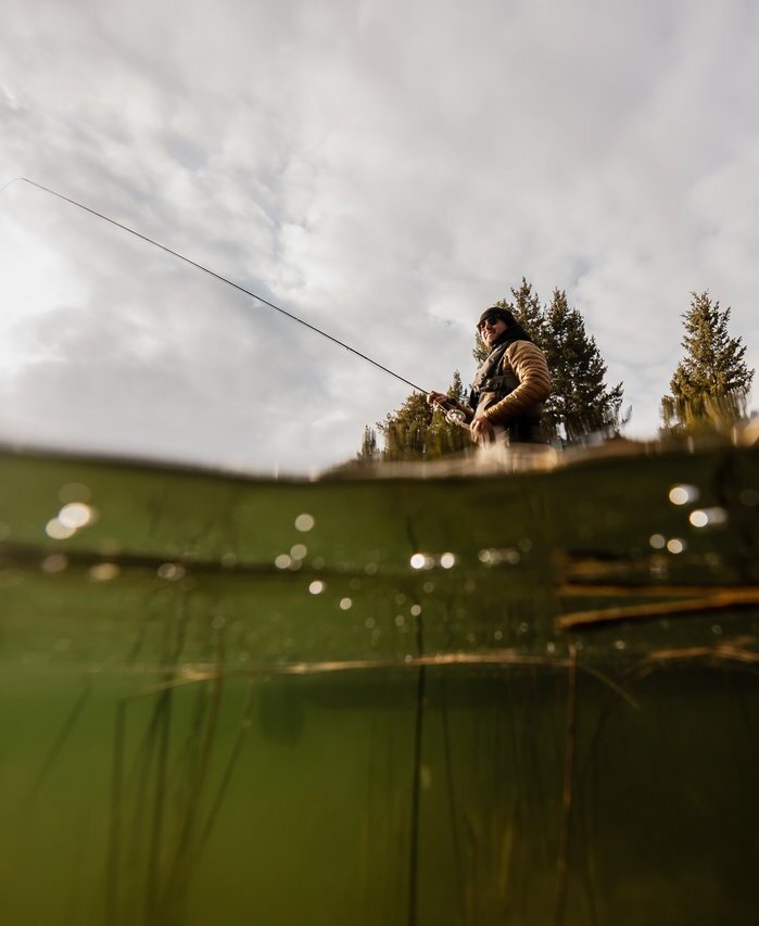 A fisher stands in a boat with a line in the water. The camera is partially submerged, showing lake weeds in the foreground.