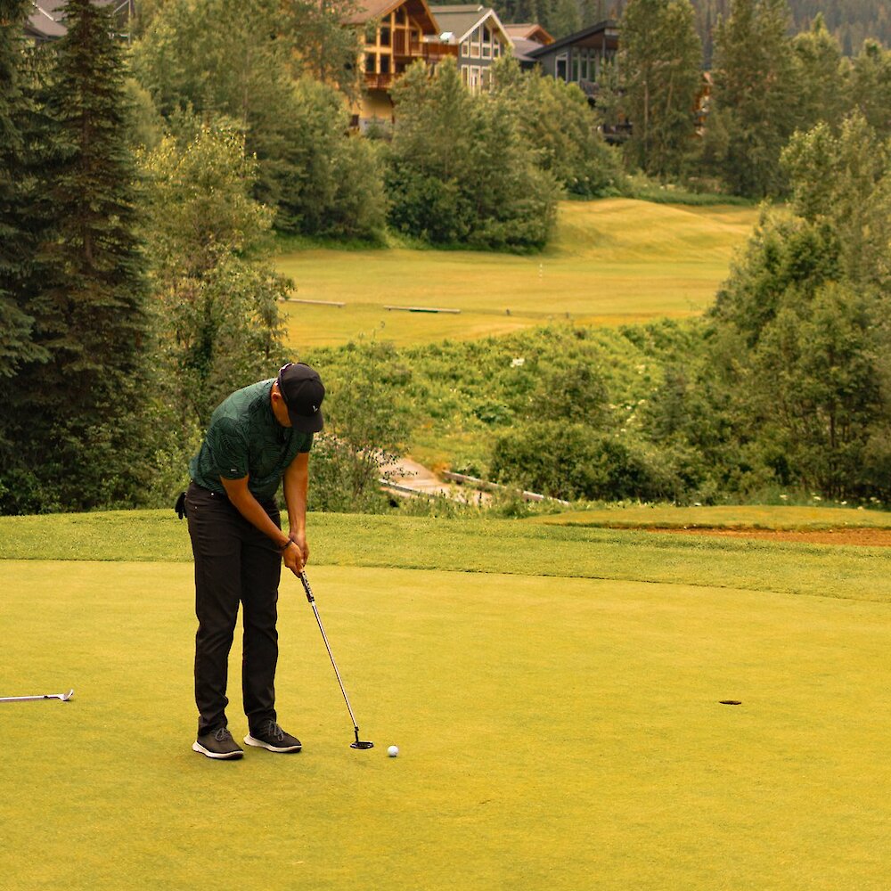 Man putting his ball with the picturesque forests and cabins in the background at Sun Peaks Golf Course in British Columbia, Canada.