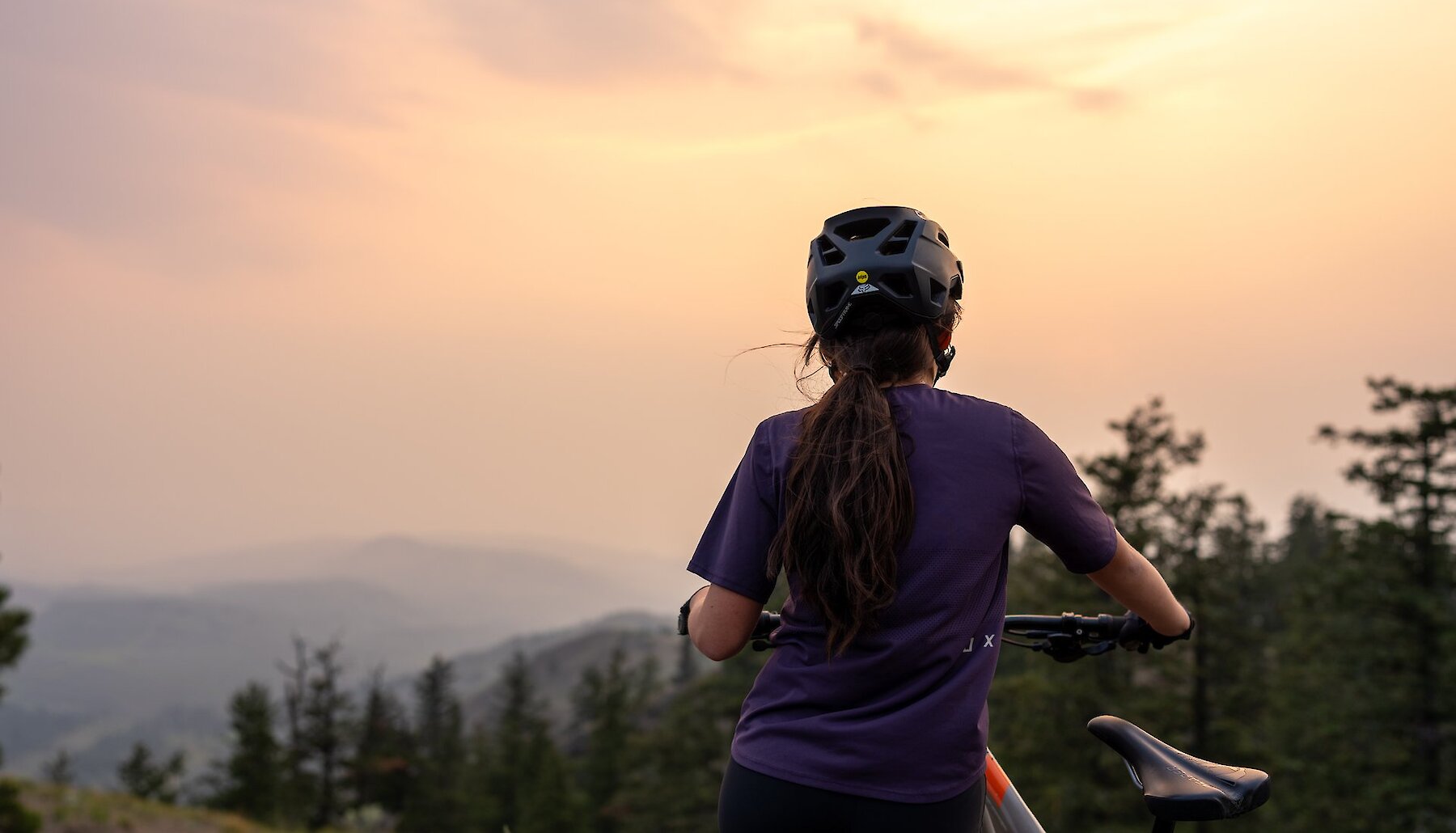 A woman holding her mountain bike with pine trees, a mountain, and sunset in the background in Kamloops, BC.