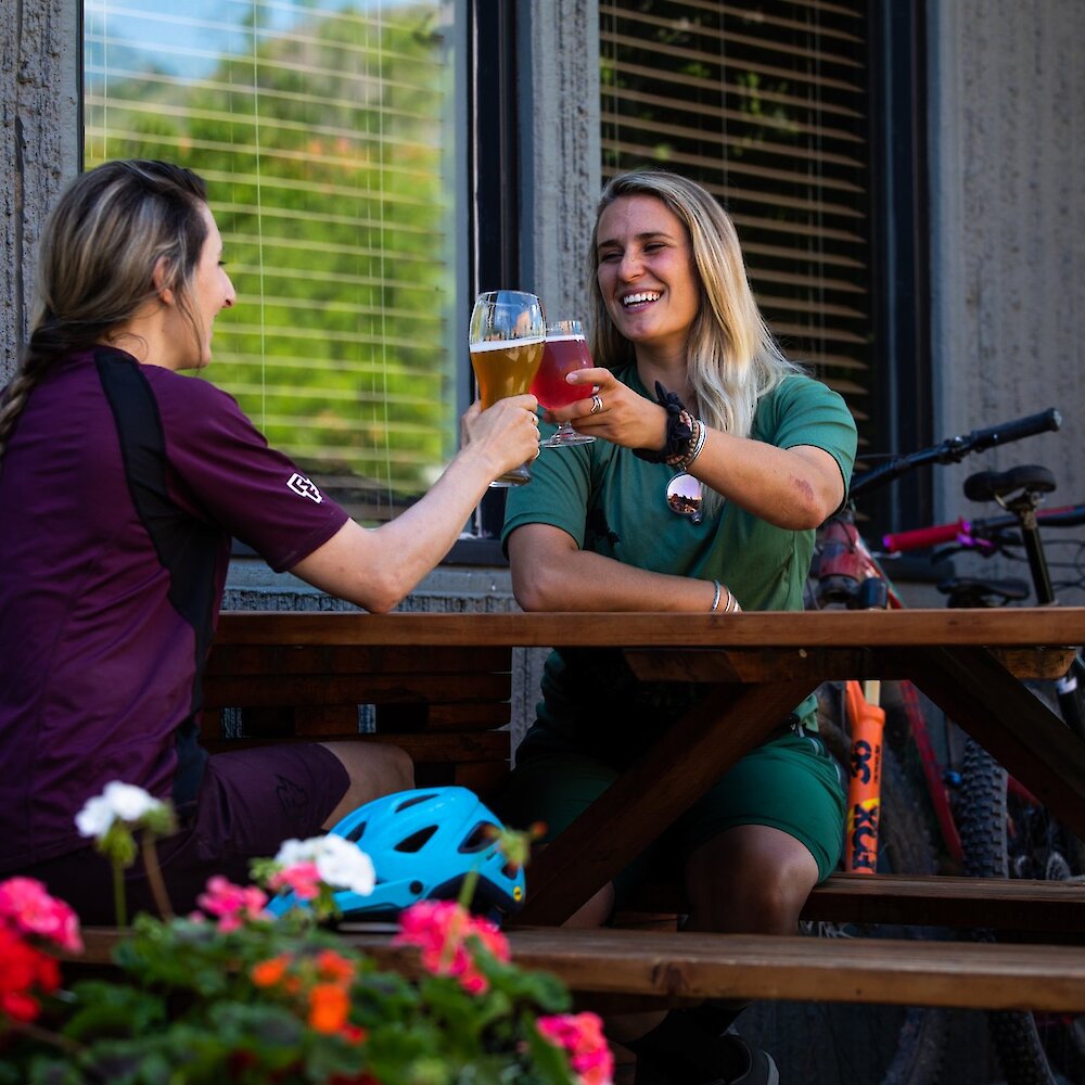 Two women cheerings their craft beer with their mountain bikes behind them on the patio at Red Collar Brewing Co in downtown Kamloops, BC.