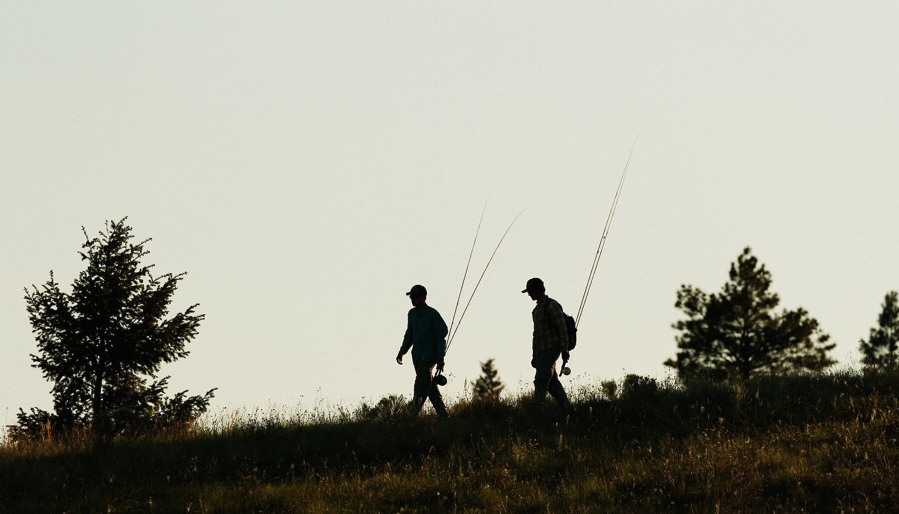 Two men holding fishing rods walking through a grassfield near Kamloops, BC.