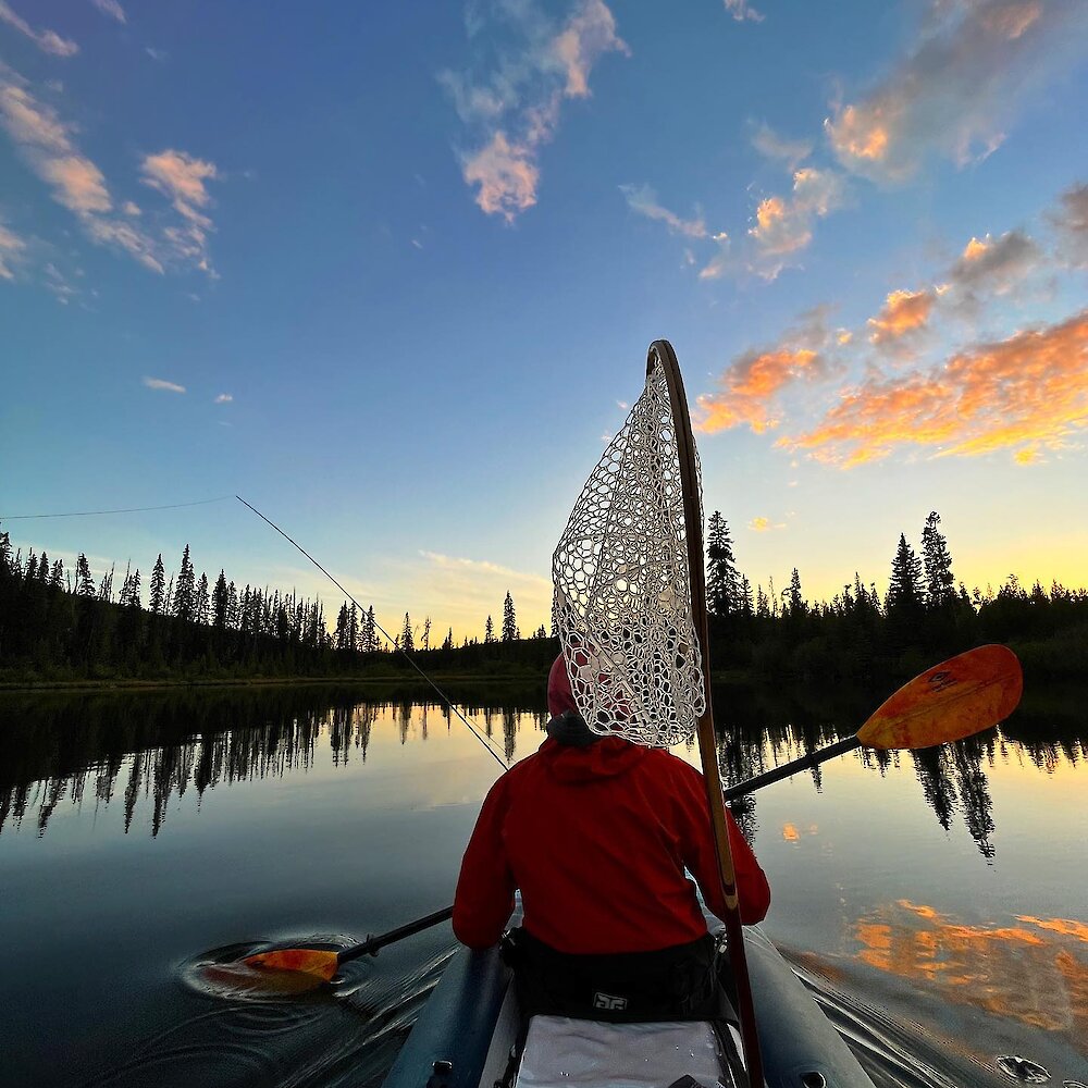 Angler paddling their boat in a Kamloops area lake with a fishing line in the water at sunset.