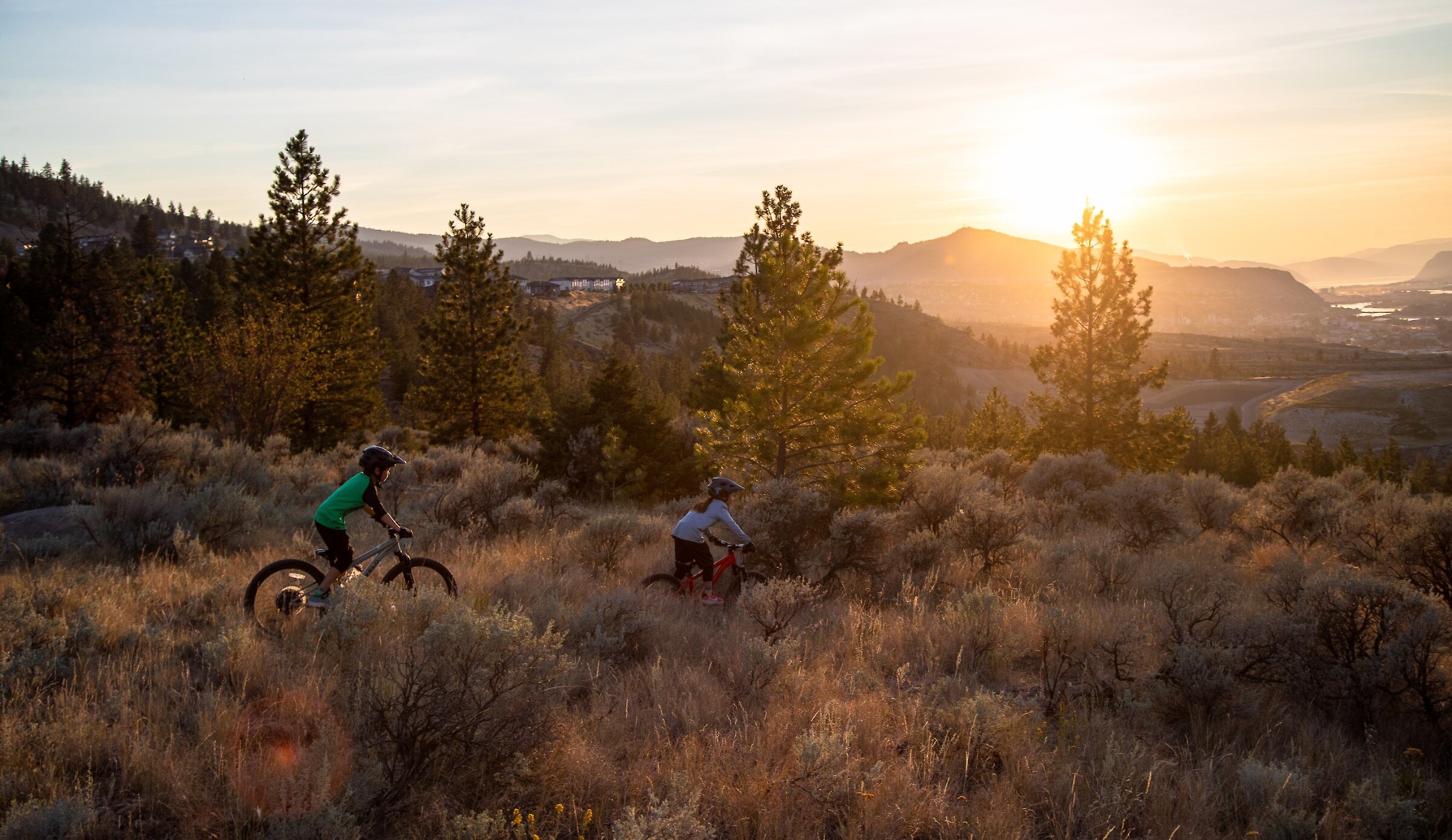 Kids mountain biking through the sagebrush at the Kamloops Bike Ranch with the sun setting over the BC mountains in the background.