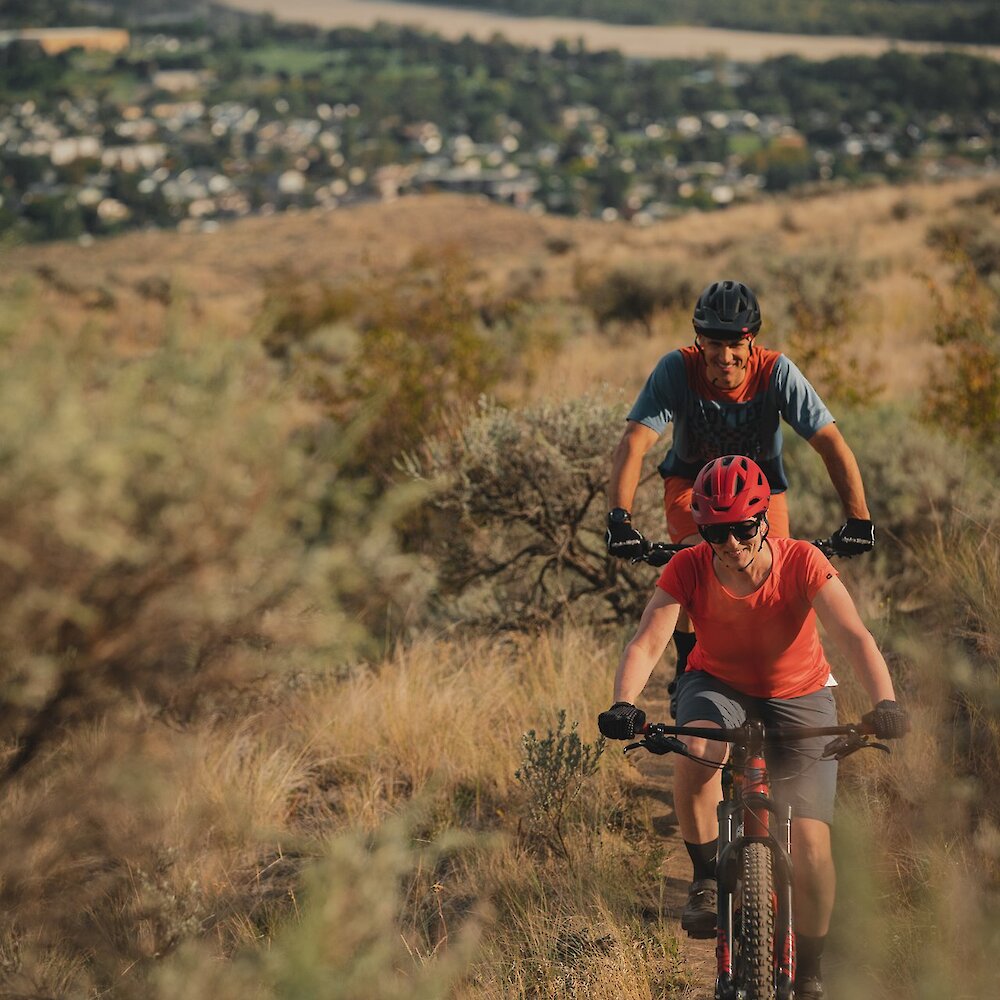 Couple biking up Lac du Bois Protected Grasslands with a view of the town of Kamloops and river in the background.