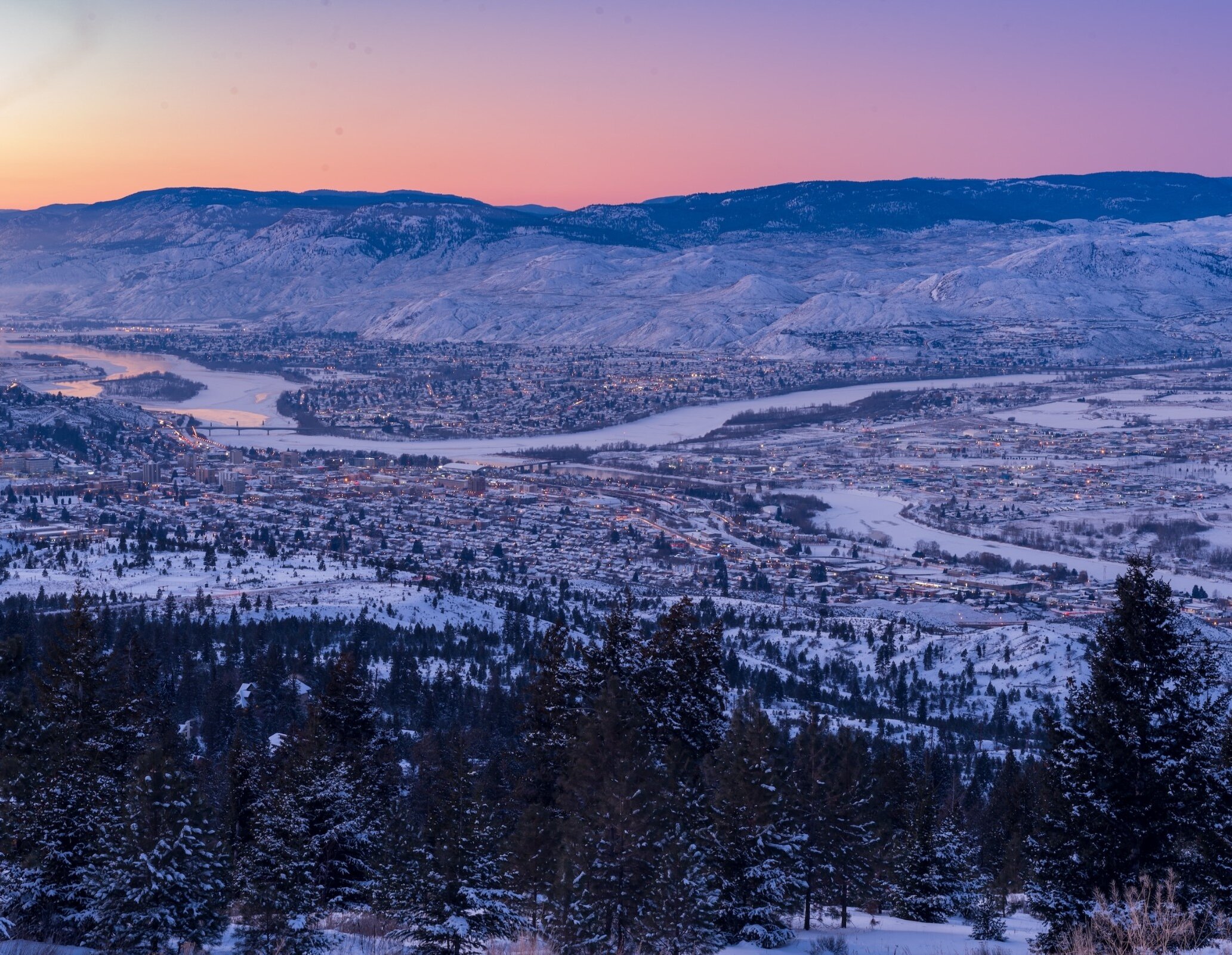 The snow covered city of Kamloops in British Columbia with the Thompson River weaving through the city and the sun setting behind the snowy mountains in the background.