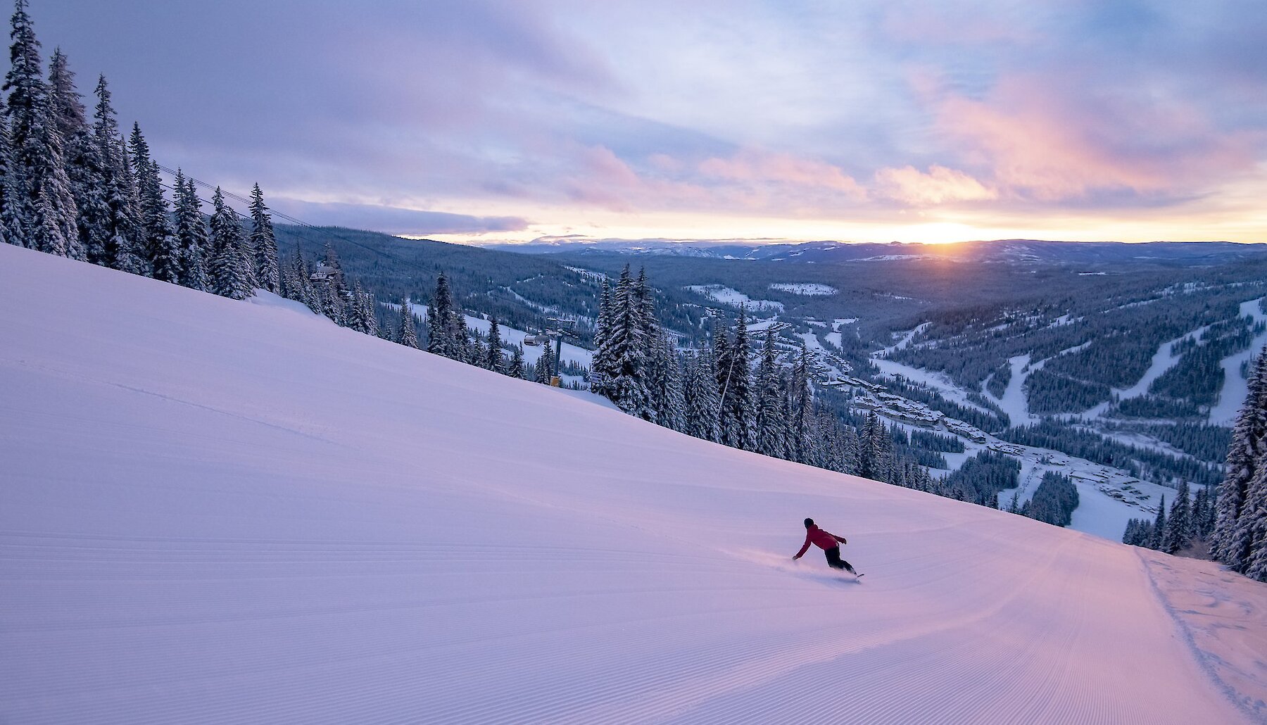 Snowboarding carving down a pristinely groomed trail at Sun Peaks Resort with a view of the mountain's ski trails and sunset in ithe background.