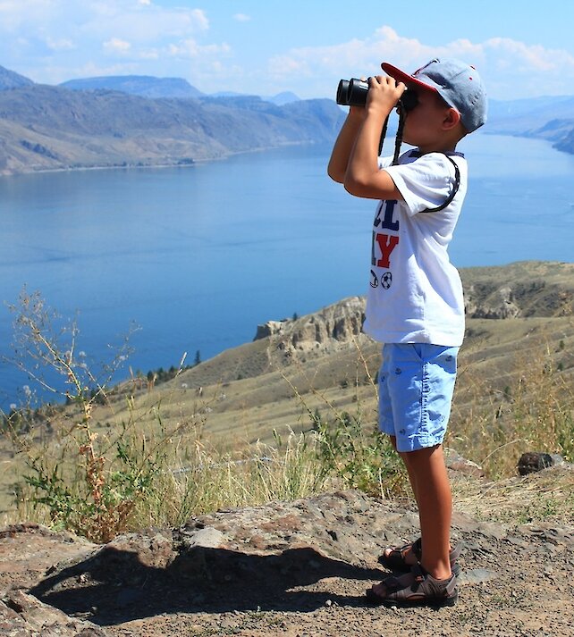 Boy looking through binoculars at the beautiful British Columbia scenery at a hiking viewpoint in Kamloops.