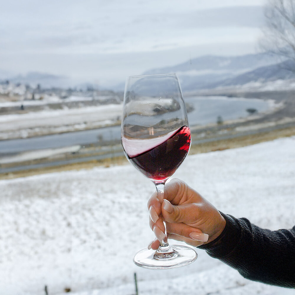 A person swirling a glasses of red wine with a snow view of the Thompson River in the background at Monte Creek Winery.