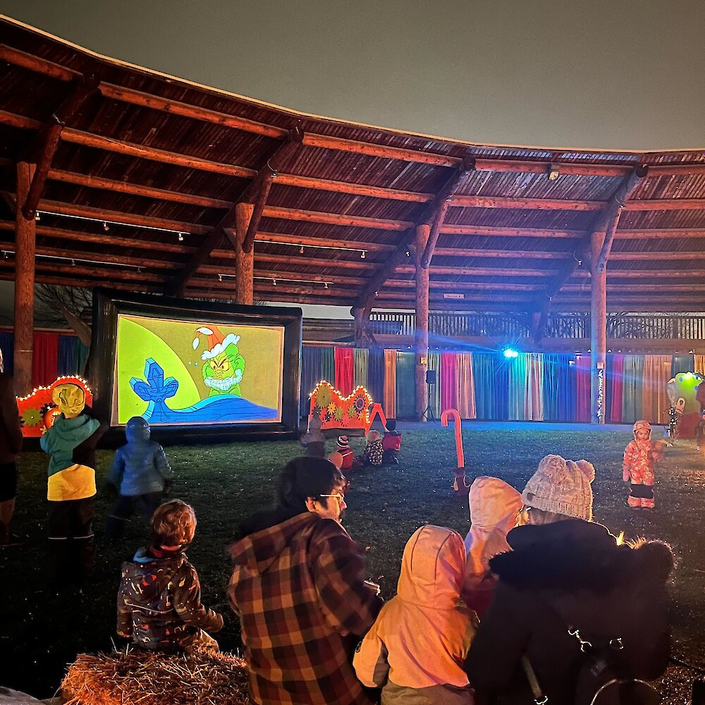 A family watching The Grinch movie on the big screen at the Who-Ville Kamloops event located at the Tk̓emlúps te Secwépemc Powwow Arbour.